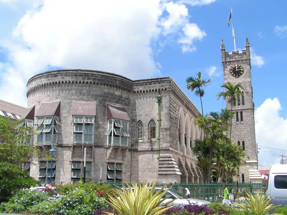 A grey stone building with a clock tower, Gothic windows, and palm trees under a blue sky with white clouds.