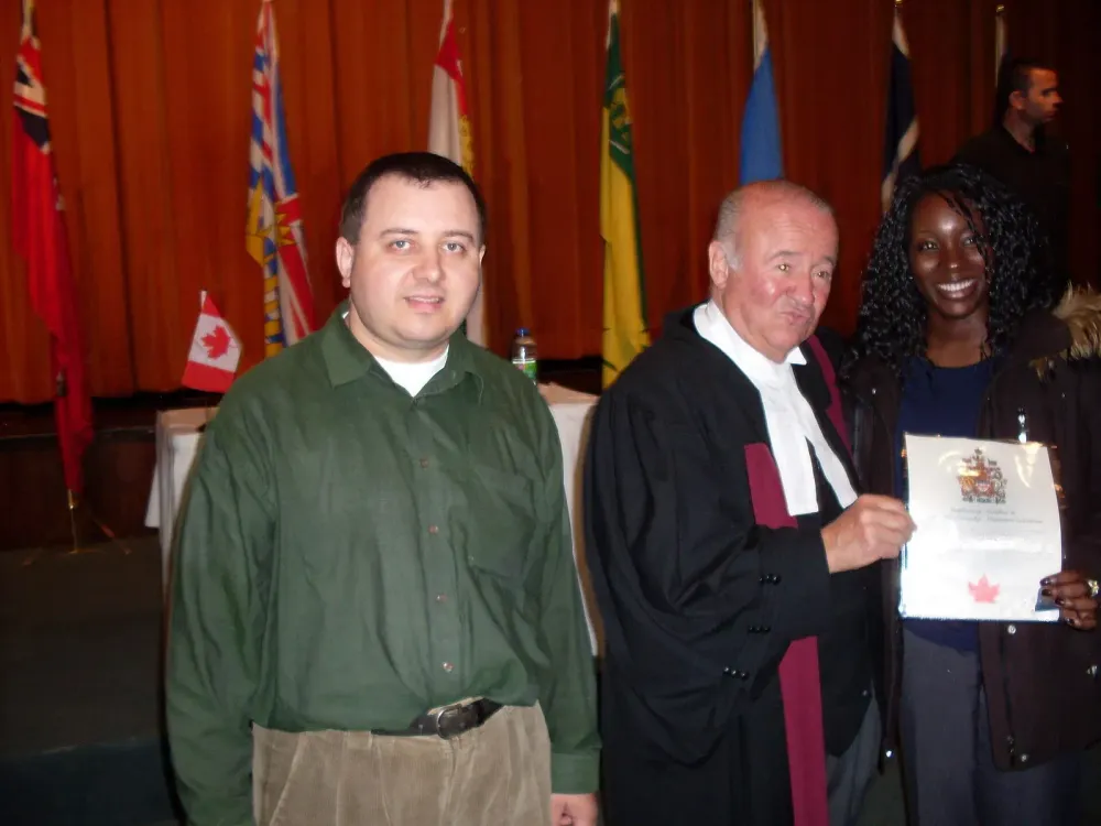 A man, a judge, and a woman holding a Canadian citizenship certificate stand in front of Canadian flags.
