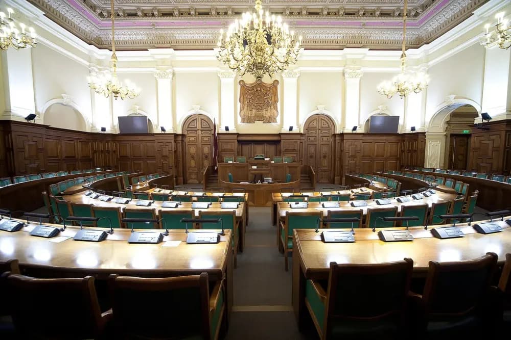 An empty legislative chamber with curved rows of wooden desks and green chairs under large crystal chandeliers.